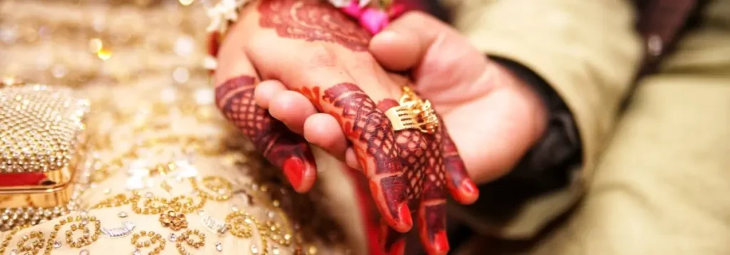 Henna-decorated hands holding each other with a gold ring, showing traditional Indian wedding attire